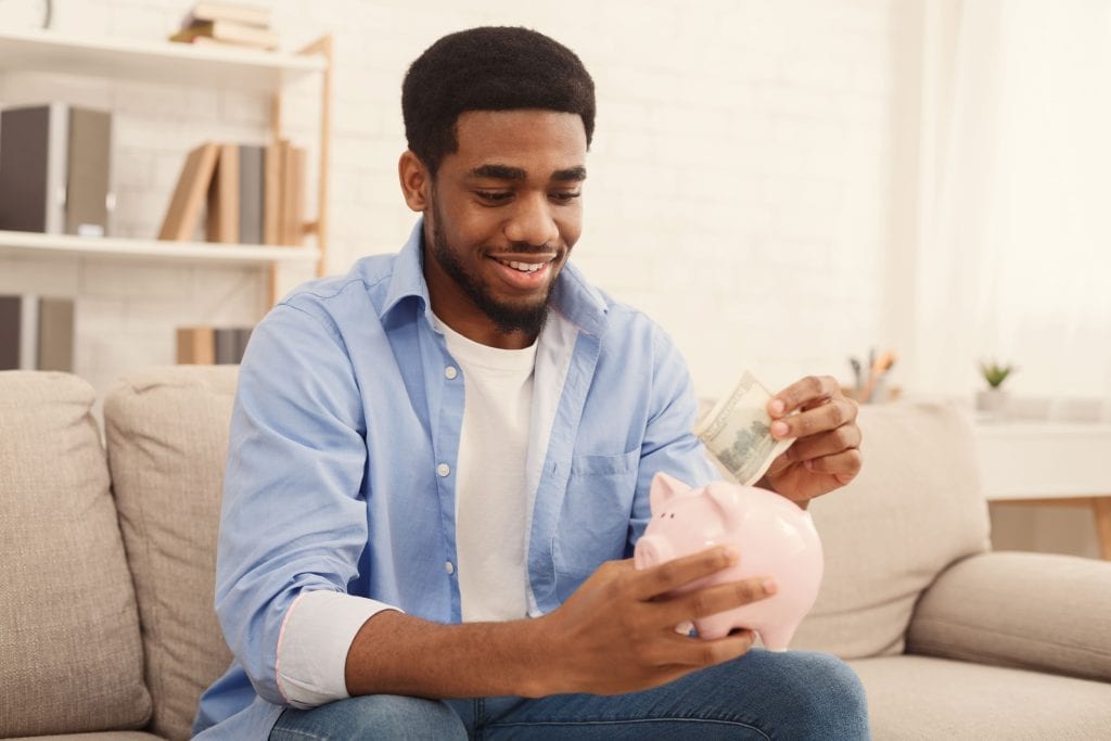 Homme de couleur souriant mettant billet dans une tirelire rose en forme de cochon