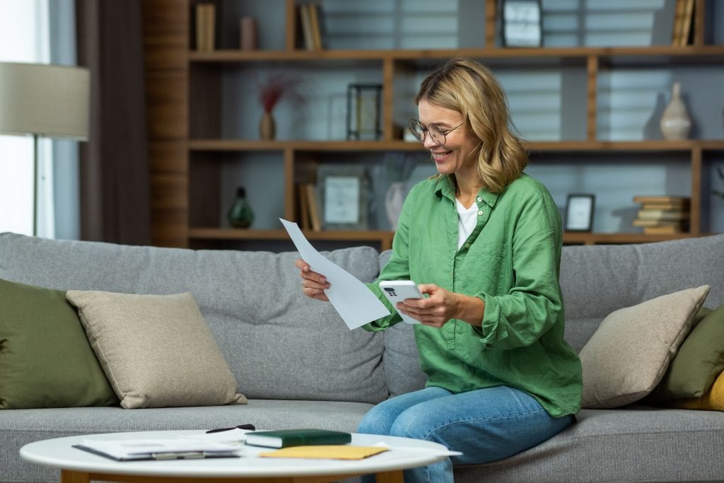 Femme blonde avec des lunettes qui regarde une facture, son téléphone à la main