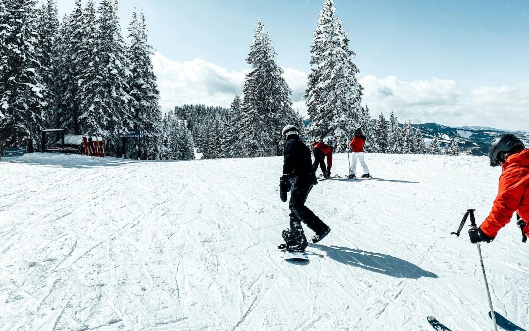 Skieurs et snowboarders sur une piste enneigée en montagne, entourés de sapins et d’un paysage hivernal, illustrant la pratique des sports d’hiver et la nécessité d’une bonne assurance avant de partir skier.